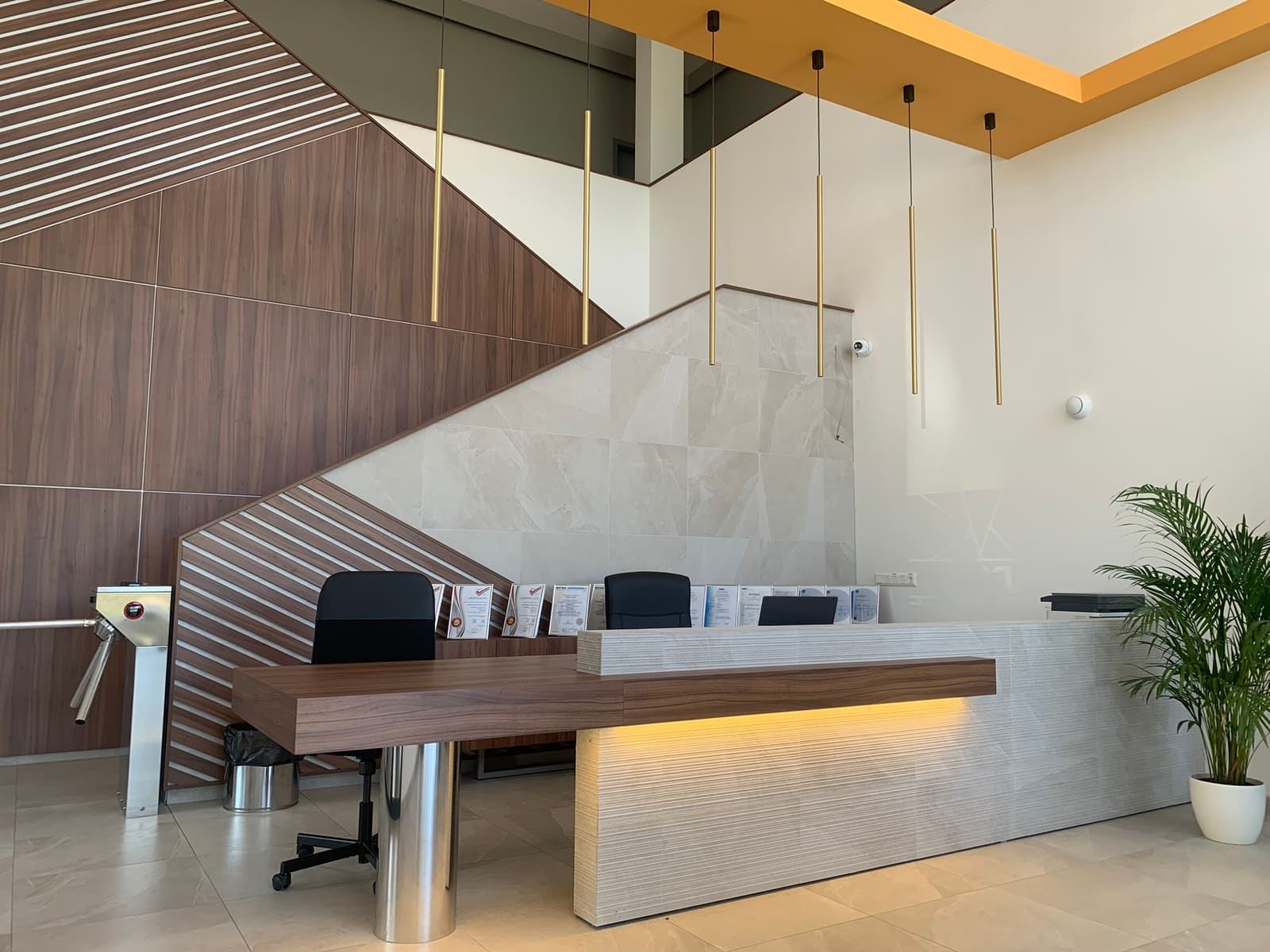 CAADEX recepció másik szögből — sárgaréz függőleges lámpák, dió csíkos akcentus, travertin hátlap, fémvázas pult, monstera