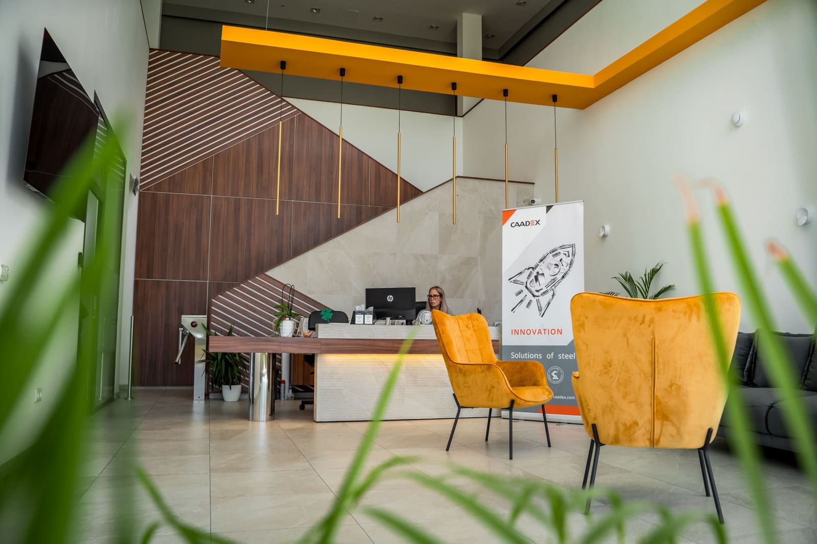 CAADEX aula a recepció felől nézve — dió csíkos akcentus, narancs LED mennyezeti keret, sárgaréz lámpák, fémvázas recepciós pult, sárga fülfotelek, CAADEX szakajtó