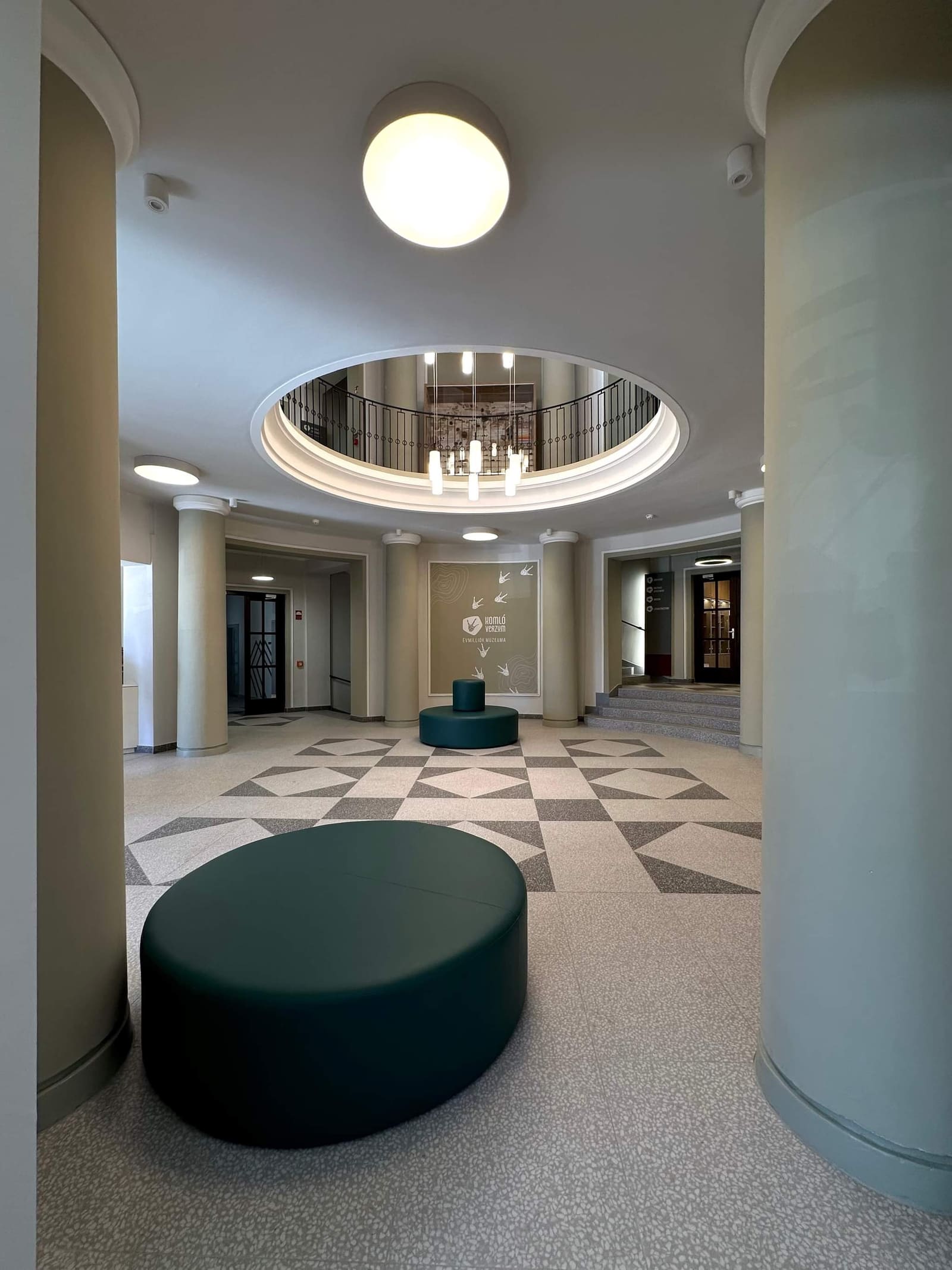 Komlói Múzeum aula — kör függesztett lámpatest a galéria nyílásában, oszlopok, terrazzo padló, sötétzöld ülőke